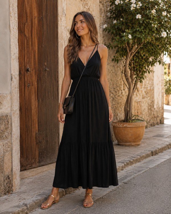 Woman wearing a black maxi dress with gold flat sandals and a black crossbody bag on a sunny Mediterranean street.